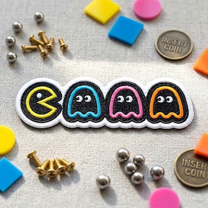 May include: A Pac-Man patch with a yellow Pac-Man character and three ghost characters in blue, pink, and orange. The patch is surrounded by a white border. Scattered around are small metal balls, screws, and colorful geometric shapes, along with two "INSERT COIN" coins.