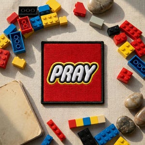 May include: A red square patch with the word "PRAY" in white letters with a yellow outline. The patch is surrounded by colorful toy building blocks and small stones. A small, aged book is in the lower left corner.