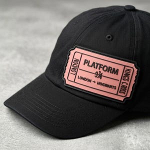 May include: A black baseball cap featuring a rectangular pink patch on the front. The patch displays black text that reads PLATFORM 9 3/4, LONDON, KINGS CROSS, and LONDON TO HOGWARTS, designed to resemble a train ticket with a black border.