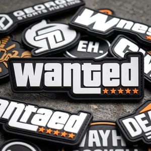 May include: A collection of black and white rubberized morale patches featuring various designs, including one prominent rectangular patch with the word wanted in bold white letters and five orange stars underneath on a black background.