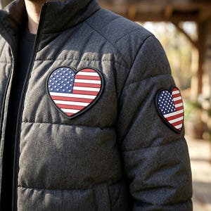 May include: A charcoal grey quilted puffer jacket featuring two heart-shaped patches with the American flag design. One patch is positioned on the upper chest area, and a smaller matching heart patch is located on the upper sleeve of the jacket.