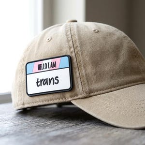 May include: A tan baseball cap featuring a rectangular patch on the front. The patch is white with a black border and displays the text HELLO I AM in black letters above the word trans in a black script font, set against a light blue and pink background.