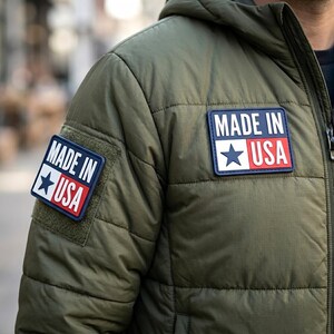 May include: An olive green quilted jacket featuring two rectangular patches with the text MADE IN USA and a blue star graphic. One patch is positioned on the upper chest and the other is on the sleeve, both displaying a white, blue, and red design.