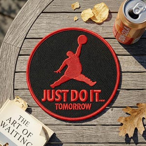 May include: A round black patch with a red embroidered figure of a person jumping with a basketball. The patch has a red border and the words "JUST DO IT... TOMORROW" in red. The patch is on a wooden surface.