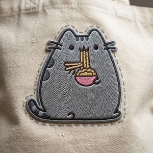 May include: A beige canvas tote bag featuring a gray embroidered cat patch. The cat is depicted eating yellow noodles from a small pink bowl with chopsticks. The embroidery includes dark gray outlines and details on the cat's body and tail.