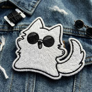 May include: Embroidered white cat patch with black sunglasses and a black outline. The patch is on a denim jacket. A spool of black thread and small sunglasses are also visible.