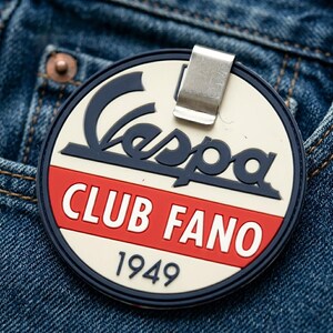 May include: A circular badge with a metal clip featuring the dark blue Vespa logo, a red horizontal stripe with the white text CLUB FANO, and the year 1949 in dark blue at the bottom, all set against a cream-colored background with a dark blue border.