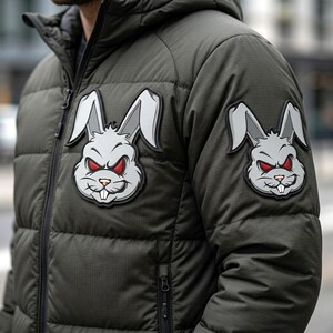 May include: An olive green puffer jacket featuring a hood and a full-length front zipper. Two identical patches are attached to the chest and sleeve, each displaying a stylized gray rabbit head with sharp features, red eyes, and visible white teeth.