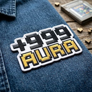 May include: Embroidered patch with the text "1999 AURA" in a pixelated font, featuring black and gold colors. The patch is on a blue denim jacket. A Game Boy cartridge and small heart-shaped objects are in the background.