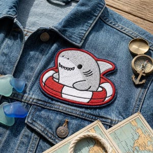 May include: Embroidered patch featuring a cartoon shark in a red and white life preserver. The patch is sewn onto a denim jacket. A compass, map, and sea glass are also visible.