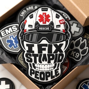 May include: A black, white, and red PVC patch featuring a skull wearing a rescue helmet with a Star of Life emblem and the text "RESCUE" and "MEDIC". Below the helmet, the skull displays the bold black text "I FIX STUPID PEOPLE" in a stylized font.