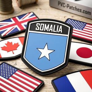 May include: A shield-shaped PVC patch featuring a light blue background with a centered white star and the text SOMALIA in white on a black header. It is surrounded by other rectangular and shield-shaped flag patches including the US, UK, and Canada.