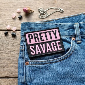 May include: A denim jeans pocket with a black and pink embroidered patch that reads "PRETTY SAVAGE." The patch is rectangular and has a pink background with black lettering. The jeans are blue and have a classic five-pocket design.