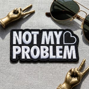 May include: A black and white embroidered patch with the text "NOT MY PROBLEM" and a heart symbol. The patch is surrounded by gold-colored peace sign sculptures and a pair of aviator sunglasses.