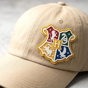 May include: A beige baseball cap featuring a centered, multi-colored rubberized crest patch. The shield-shaped emblem is divided into four quadrants displaying a white lion, snake, badger, and eagle, with a central yellow letter H on a gold background.