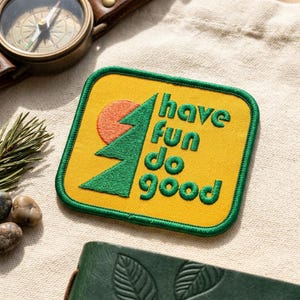 May include: Yellow embroidered patch with the words "have fun do good" in green. The patch features a green tree and a red sun design. A compass and a leather strap are visible in the background.