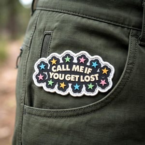 May include: A black, cloud-shaped patch with a white border featuring the text CALL ME IF YOU GET LOST in cream-colored letters. The patch is decorated with small, colorful stars in shades of pink, yellow, light blue, and green, attached to olive green fabric.