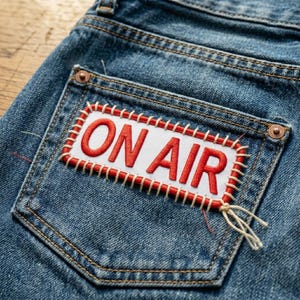 May include: Close-up of blue denim jeans with a back pocket featuring a white patch. The patch has the words "ON AIR" embroidered in red, outlined with a red border and white stitching. The jeans have classic stitching details.