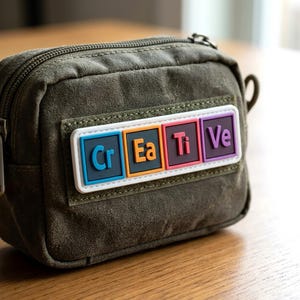 May include: An olive green canvas zippered pouch featuring a rectangular patch with the word "Creative" spelled out in colorful, individual square tiles. Each letter tile has a distinct border and background color, including blue, orange, red, and purple.
