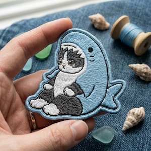 May include: An embroidered patch featuring a black and white cat wearing a light blue shark costume. The cat has large yellow eyes and is positioned inside the shark's open mouth. The patch has a detailed stitched border and a soft fabric texture.