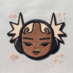 May include: Embroidered patch featuring a stylized face with headphones, a nose ring, and starburst accents. The face is brown with black details and light brown hair. Additional star and sound wave designs are embroidered around the main image.