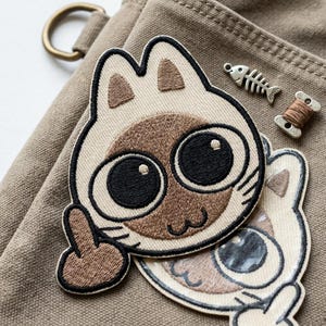 May include: Embroidered patch featuring a Siamese cat with large black eyes and a brown face, giving the middle finger. A small fish-shaped charm and a spool of brown thread are also visible. The patch is on a tan fabric.