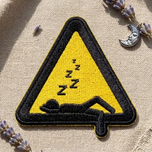 May include: Embroidered patch featuring a yellow triangle with a black border. Inside, a black silhouette of a person sleeping is depicted, with 'Z's above. The patch is on a neutral fabric background, with lavender sprigs and a crescent moon charm.