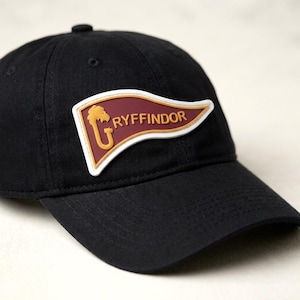 May include: A black baseball cap featuring a pennant-shaped patch on the front. The patch is maroon with a white border and displays the text GRYFFINDOR in gold lettering, accompanied by a gold lion emblem integrated into the letter G.