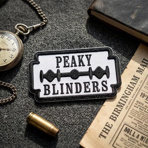 May include: A rectangular patch with the words "PEAKY BLINDERS" in black, set against a white background, and a black border. The design includes a stylized razor blade. A vintage pocket watch and a bullet casing are also visible.
