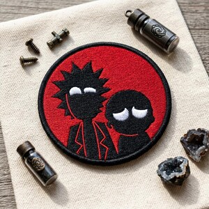 May include: A round, embroidered patch featuring black silhouettes of two cartoon characters on a red background. The patch is surrounded by small metal vials, screws, and geodes on a beige fabric surface.