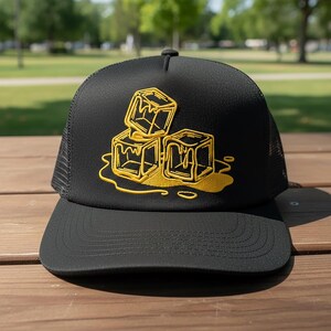 May include: Black trucker hat with a mesh back and a curved brim. The front of the hat features a yellow embroidered design of three melting ice cubes. The hat is sitting on a wooden surface.