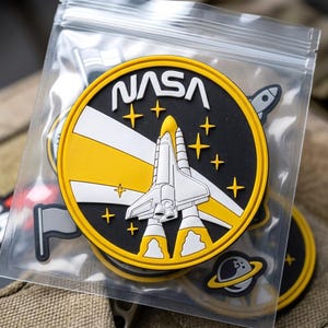 May include: A circular NASA patch in a clear plastic bag. The patch features a white space shuttle launching against a black background with yellow stars and a yellow light beam. The text NASA is printed in white at the top of the circular design.