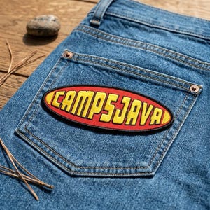 May include: Close-up of blue denim jeans with a back pocket. A red and yellow embroidered patch that reads "CAMPSJAVA" is sewn onto the pocket. The patch is oval-shaped. The jeans are on a wooden surface.