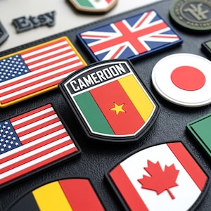 May include: A collection of rectangular and shield-shaped rubberized patches featuring various national flags, including the United States, United Kingdom, Japan, Canada, and a shield-shaped patch with the text CAMEROON and the national flag.