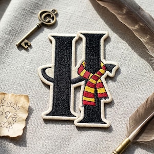 May include: Embroidered patch featuring the letter "H" in black with a cream border, adorned with a red and yellow striped scarf. A vintage key, feather, and parchment with symbols are also present, suggesting a theme of fantasy or magic.