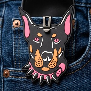 May include: A black rubberized Doberman dog head accessory featuring pink inner ears, orange muzzle markings, and a pink spiked collar with black spikes. The item includes a silver-toned metal clip attached to the top for securing onto fabric.