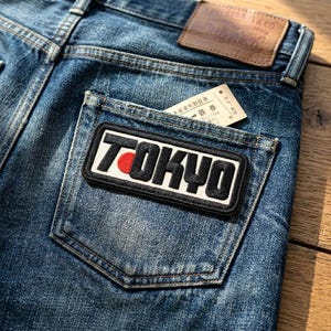 May include: Close-up of blue denim jeans with a TOKYO patch on the back pocket. The patch is rectangular with black lettering, a red circle, and a white background. A small paper ticket is in the pocket.
