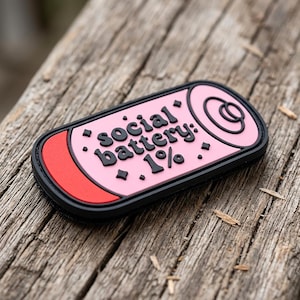 May include: A rectangular patch with rounded corners featuring a pink background, a black border, and a red accent bar on the left. The text "social battery: 1%" is printed in black, accompanied by small black diamond-shaped icons and a circular graphic.