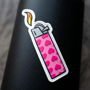 May include: A rectangular sticker featuring a pink lighter with a repeating pattern of dark pink hearts. The lighter has a grey top mechanism and a stylized flame with orange and yellow colors, all outlined in a thick white border.