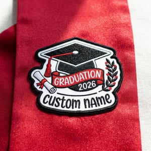 May include: A graduation patch with a black mortarboard, a red scroll, and the text "Graduation 2026 Custom Name." The patch is on a red satin background. The design includes a laurel wreath.
