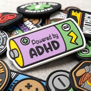 May include: A rectangular rubber patch shaped like a battery with a light purple center and green ends. The text "Powered by ADHD" is printed in black letters. A yellow lightning bolt icon and a plus sign are visible on the battery surface.