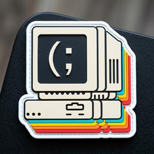 May include: A rectangular rubber patch featuring a beige vintage computer monitor and base unit. The screen displays a black background with a white winking emoticon. The design includes a layered, colorful shadow effect in teal, yellow, and red.
