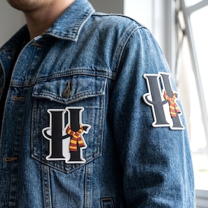May include: A blue denim jacket featuring two identical patches. Each patch displays a large black letter H with a white outline, wrapped in a red and yellow striped scarf. The patches are positioned on the chest pocket and the upper sleeve.