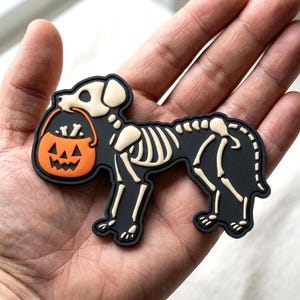 May include: A black rubberized patch shaped like a dog skeleton with cream-colored bones. The dog is holding an orange jack-o'-lantern bucket in its mouth, which contains small white bone shapes. The design features a flat, matte finish.