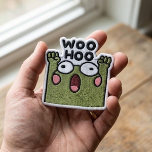 May include: An embroidered patch featuring a green square character with raised arms, wide eyes, and a surprised expression with pink cheeks. The text WOO HOO is stitched in black above the character, all outlined in white thread.