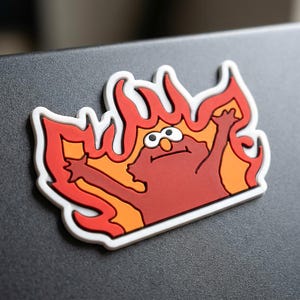 May include: A rubberized magnet featuring a red character with wide eyes and raised arms, positioned in front of stylized orange and red flames. The design is outlined in a thick white border, creating a high-contrast graphic representation.