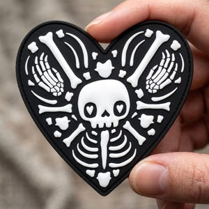 May include: A heart-shaped patch featuring a black background with a white graphic design of a central skull with heart-shaped eye sockets, surrounded by skeletal hands, rib cage elements, and various scattered bone shapes in a symmetrical layout.