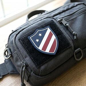 May include: A charcoal gray tactical sling bag featuring a shield-shaped morale patch on the front. The patch displays a blue field with two white stars and red and white vertical stripes. The bag includes zippered compartments and metal D-rings.