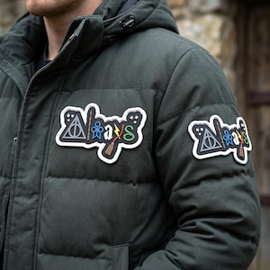 May include: A dark olive green puffer jacket featuring two identical patches with the word Always in a stylized font. The design incorporates a triangular symbol, a wand, a broomstick, and a lightning bolt, all outlined in white on a black base.