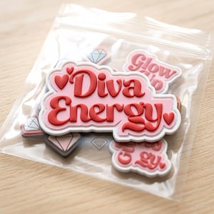 May include: A clear plastic bag containing multiple rubber charms. The primary charm is a pink, cloud-shaped piece with the red text "Diva Energy" and small red hearts. Other charms include diamond shapes and additional "Diva Energy" text pieces.